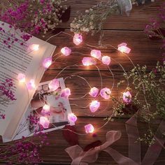 an open book with pink flowers and fairy lights around it on top of a wooden table