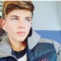 a young man sitting on a bus wearing a jacket and looking at the camera with blue eyes