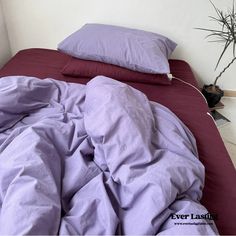 an unmade bed with purple sheets and pillows on top of it next to a potted plant