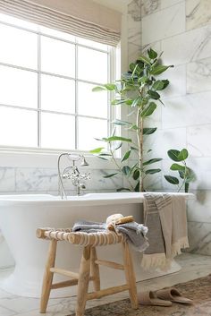 a white bath tub sitting under a window next to a wooden stool and potted plant