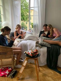 four women sitting on a bed in a room with white walls and wooden flooring