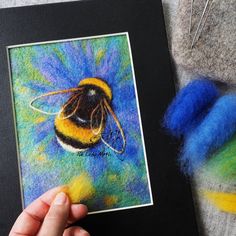 a hand holding up a card with a painting of a bee on it and some yarn next to it