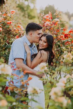 a man and woman standing in the middle of flowers with their arms around each other