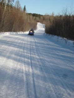 two people on snowmobiles going down a snowy road with trees in the background