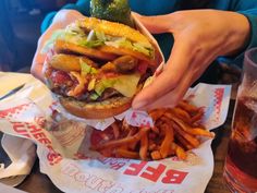 a person holding up a large hamburger and french fries in front of a glass of beer