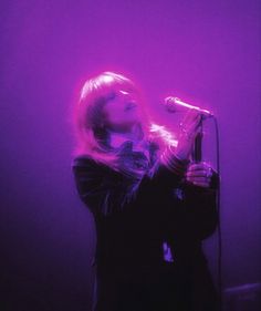 a woman singing into a microphone while standing in front of a purple wall with lights