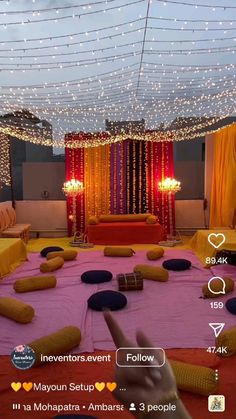 a room decorated with yellow and purple pillows on the floor, lights strung over the ceiling