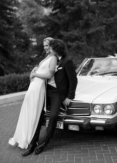 black and white photo of bride and groom leaning on car with trees in the background