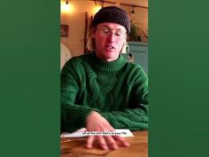 a man sitting at a table wearing a hat and green sweater with writing on it