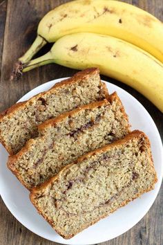 three slices of banana bread on a white plate next to two bananas and a wooden table