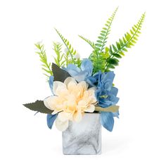 a white vase with blue and yellow flowers in it on a white table top next to some green leaves