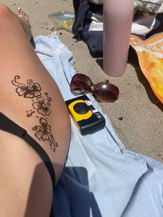 a woman laying on top of a towel next to a bottle and sunglasses with a tattoo on her stomach