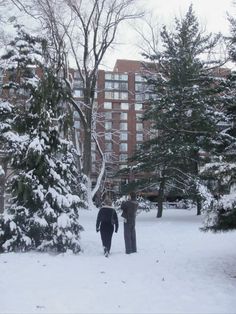 two people are walking through the snow in front of some trees and apartment buildings on a snowy day