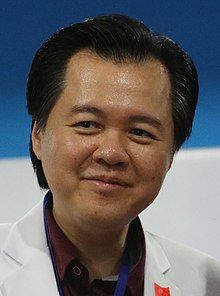 a man in a white lab coat smiles at the camera while wearing a medal around his neck