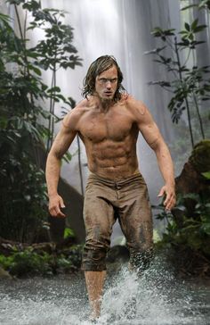 a man with no shirt is walking through the water in front of a waterfall and trees