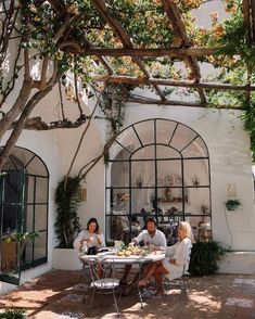 three people sitting at a table under a pergolated trellis in front of a house