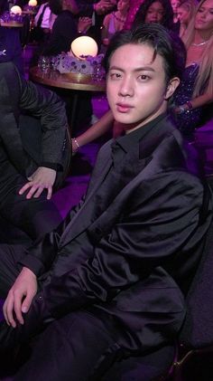 a young man in a black suit sitting at a table with other people around him