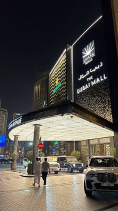 two people walking in front of a building at night with cars parked on the street