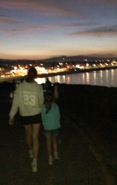 a woman and child are standing on the edge of a hill at night with city lights in the background