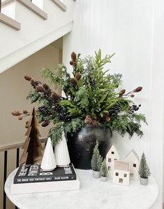 a white table topped with a black vase filled with pine cones and greenery next to a stair case