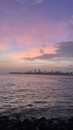 the sky is purple and pink as it sets over the ocean with buildings in the distance