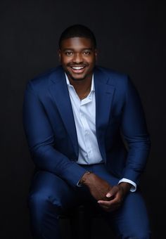 a man in a blue suit and white shirt is sitting on a stool smiling at the camera