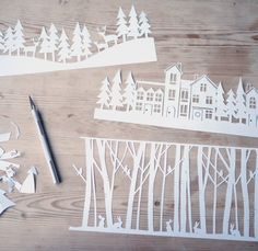 cut out paper trees and houses on a wooden surface with a pen next to them