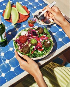 a person holding a plate of food with meatballs and vegetables on the table next to them