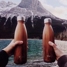 two people holding brown water bottles in front of a mountain lake with snow capped peaks