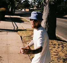 a man in white shirt and blue hat standing on sidewalk with skateboard near tree