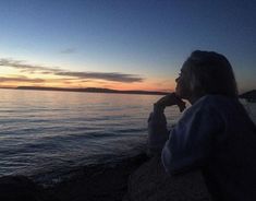 a woman sitting on top of a rock next to the ocean at sunset with her hand under her chin