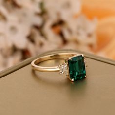 an emerald and diamond ring sitting on top of a gold box with flowers in the background