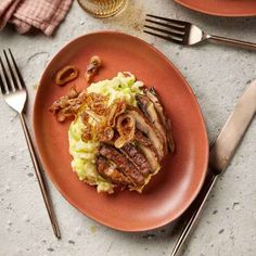 an orange plate topped with mashed potatoes covered in onions and sliced mushrooms next to silverware