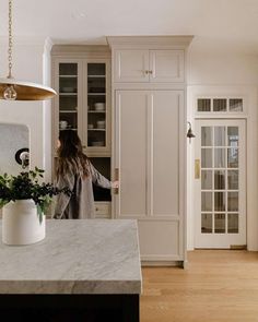 a woman standing in a kitchen next to a white cabinet and counter top with plants on it