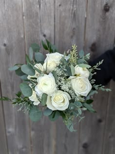 a bouquet of white roses and greenery in front of a wooden fence with someone holding it