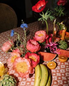 a table topped with flowers and fruit on top of a brown table cloth next to candles