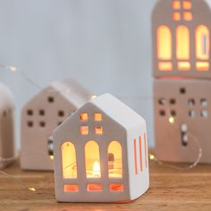 a small white house shaped candle holder on a wooden table with some lights around it