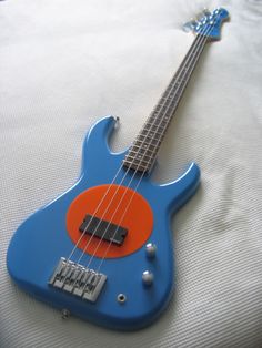 a blue and orange bass guitar laying on top of a white bed sheet with an orange dot in the middle