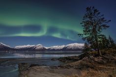 the northern lights shine brightly over snow covered mountains and trees near a body of water