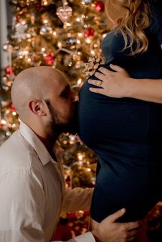 a pregnant woman kissing her husband's belly in front of a decorated christmas tree