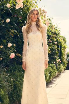 a woman in a white dress standing on a sidewalk next to flowers and greenery