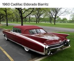 an old red car parked in a parking lot next to some trees and green grass