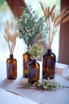 three brown glass bottles filled with flowers and greenery on top of a white table cloth