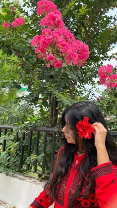 a woman with long black hair wearing a red shirt and flower in her hair standing next to a fence