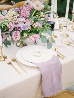 the table is set with white plates and goldware, purple flowers, and greenery