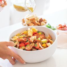 a person is pouring dressing into a bowl with vegetables and bread on the table next to them