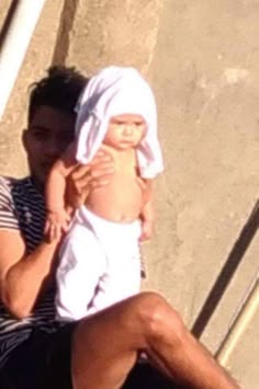 a man holding a baby wearing a white towel on his head and sitting next to stairs