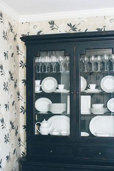 a black china cabinet with glass doors and dishes on the top, in front of a floral wallpapered wall