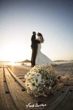 a bride and groom standing on a wooden deck with flowers in front of the sun