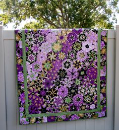 a quilt hanging on the side of a fence with purple flowers and green trim around it